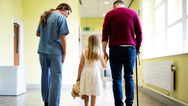 Man with a cane holding a girl’s hand while walking in a corridor and talking to a nurse
